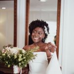 Smiling bride with natural curly hair and white floral accessories in off-shoulder wedding dress holding bouquet