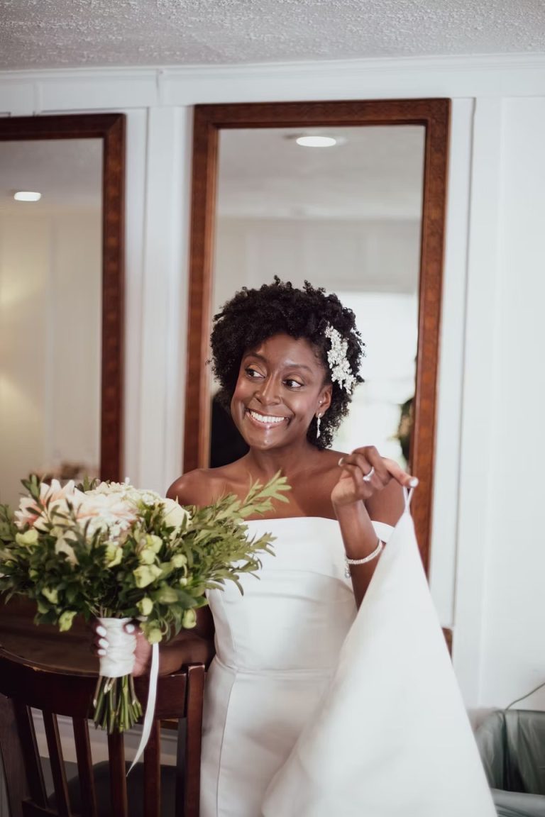 Smiling bride with natural curly hair and white floral accessories in off-shoulder wedding dress holding bouquet