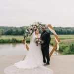 Bride in lace gown with long veil and groom in black suit standing before geometric wedding arch by pond