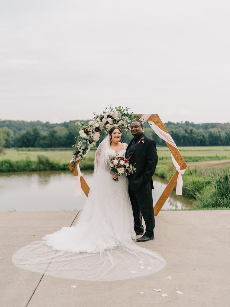 Bride in lace gown with long veil and groom in black suit standing before geometric wedding arch by pond