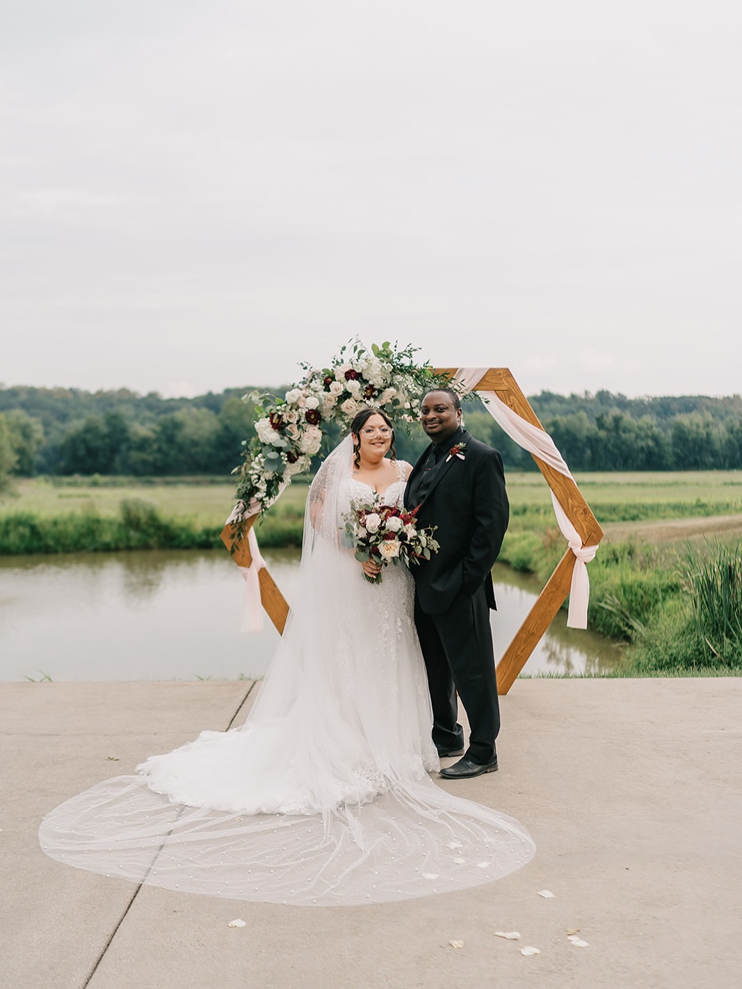 Bride in lace gown with long veil and groom in black suit standing before geometric wedding arch by pond