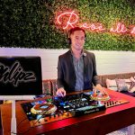 DJ in blazer and checkered shirt performing at outdoor venue with neon "Rosé all day" sign and greenery wall backdrop
