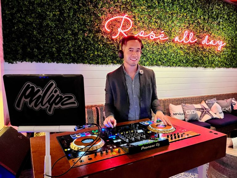 DJ in blazer and checkered shirt performing at outdoor venue with neon "Rosé all day" sign and greenery wall backdrop