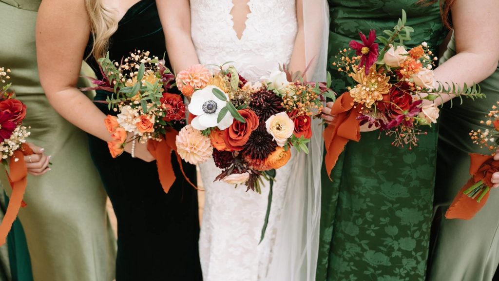 Bride and bridesmaids holding vibrant fall bouquets with dahlias, anemones, and roses in orange, burgundy, and cream tones