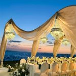 Elegant beachfront wedding reception with draped fabric canopy, crystal chandeliers, white floral centerpieces, and ocean sunset backdrop