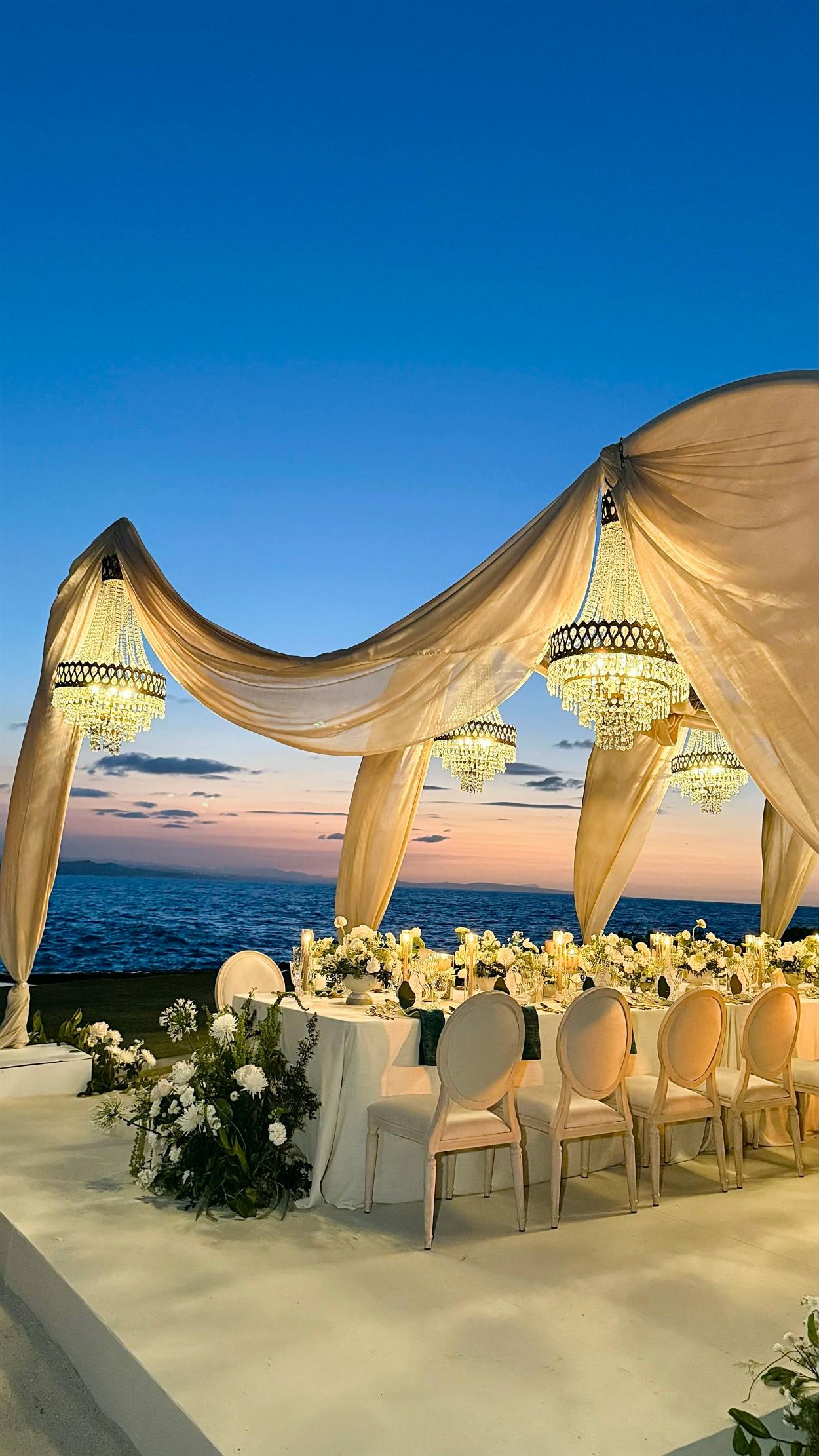 Elegant beachfront wedding reception with draped fabric canopy, crystal chandeliers, white floral centerpieces, and ocean sunset backdrop
