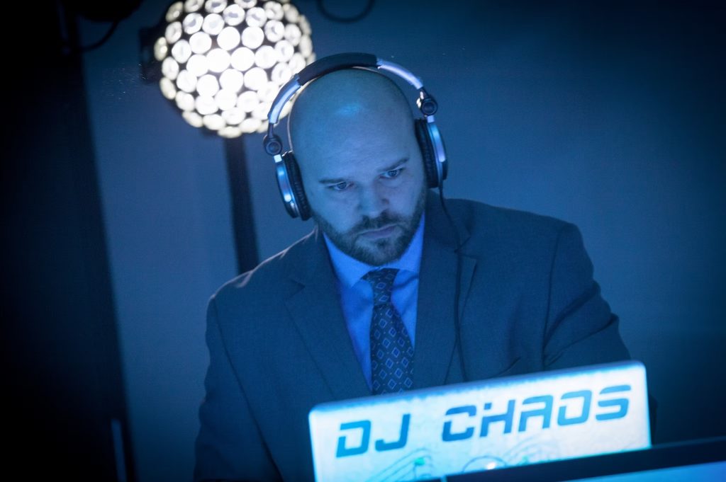 Wedding DJ in suit and headphones performing at event with illuminated DJ Chaos booth and disco ball