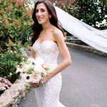 Bride in lace strapless gown with veil holding pink and white bouquet in garden setting