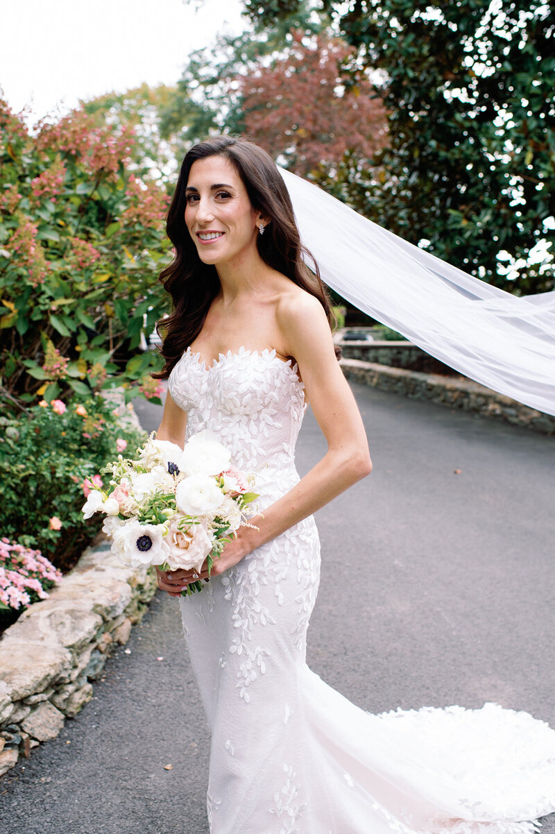 Bride in lace strapless gown with veil holding pink and white bouquet in garden setting