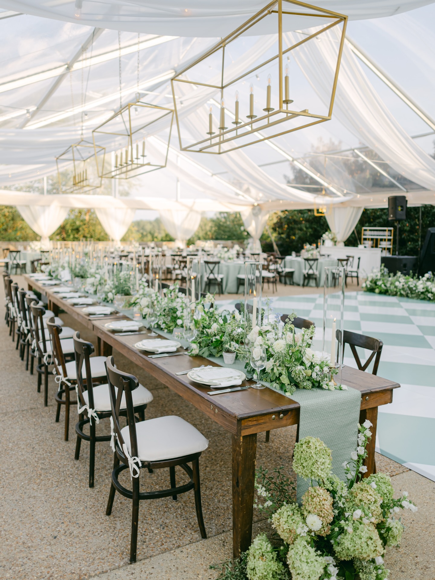 Elegant tented wedding reception with long farm tables, geometric gold chandeliers, and lush greenery centerpieces