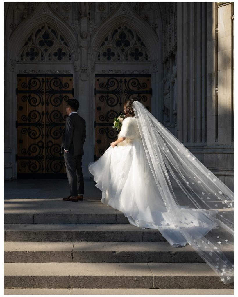 Bride in flowing white gown with long cathedral veil ascending church steps with groom before ornate Gothic doorway