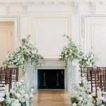 Indoor wedding ceremony setup in elegant ballroom with fireplace, floral-adorned aisle, and brown chiavari chairs
