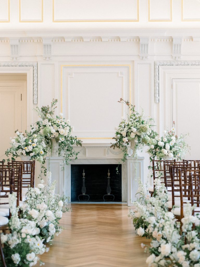 Indoor wedding ceremony setup in elegant ballroom with fireplace, floral-adorned aisle, and brown chiavari chairs