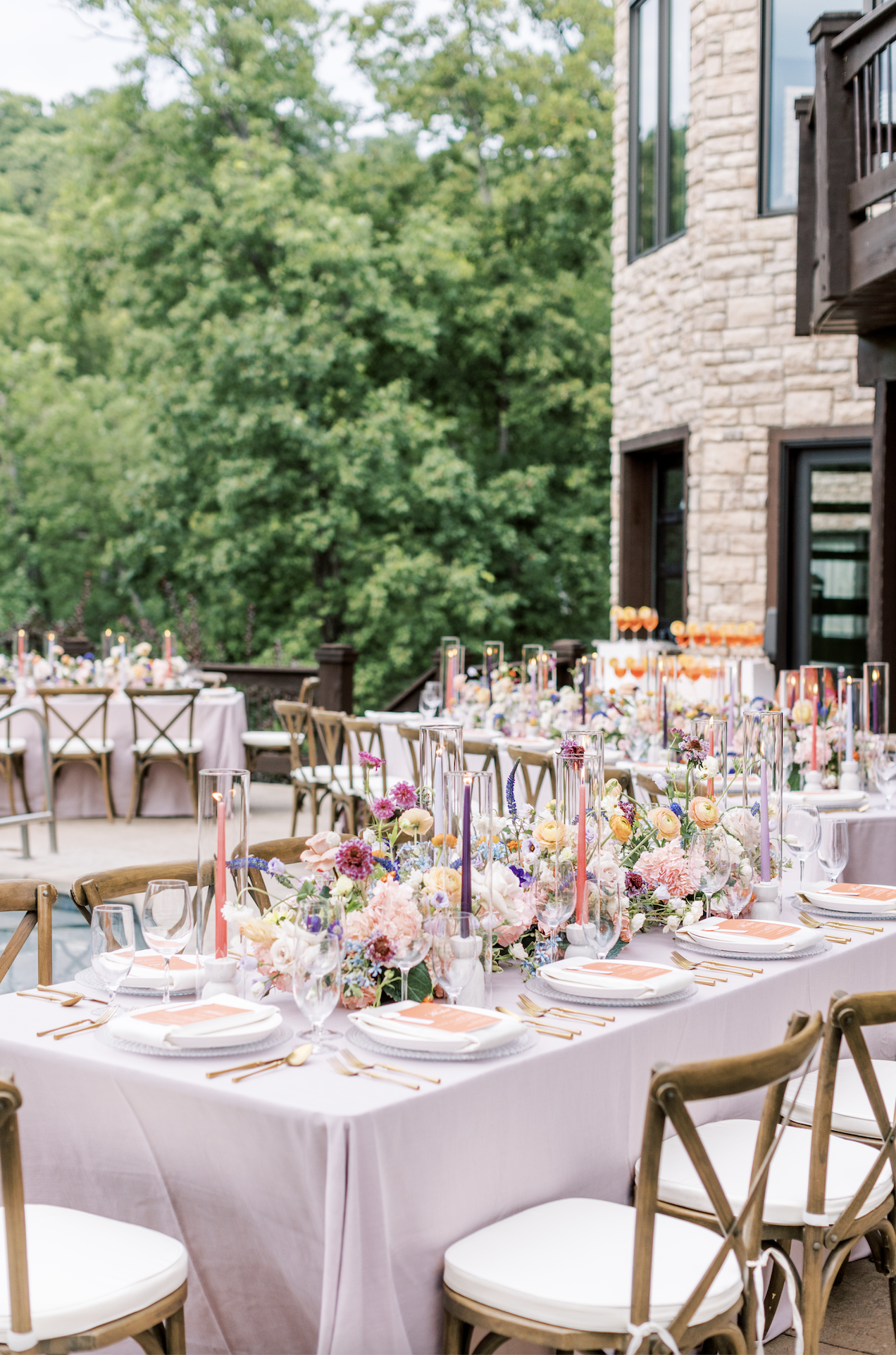 Outdoor wedding reception patio with lavender linens, wooden cross-back chairs, and colorful floral centerpieces with taper candles
