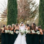 Bride in lace wedding gown with bridesmaids in black dresses holding white bouquets outdoors among evergreen trees