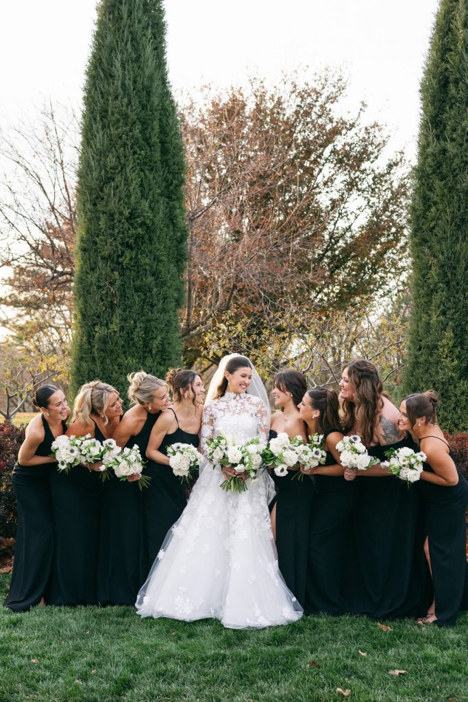 Bride in lace wedding gown with bridesmaids in black dresses holding white bouquets outdoors among evergreen trees