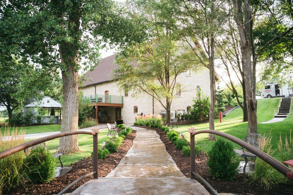 Stone building wedding venue with tree-lined walkway and green railing, surrounded by landscaped gardens