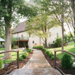 Stone building wedding venue with tree-lined walkway and green railing, surrounded by landscaped gardens