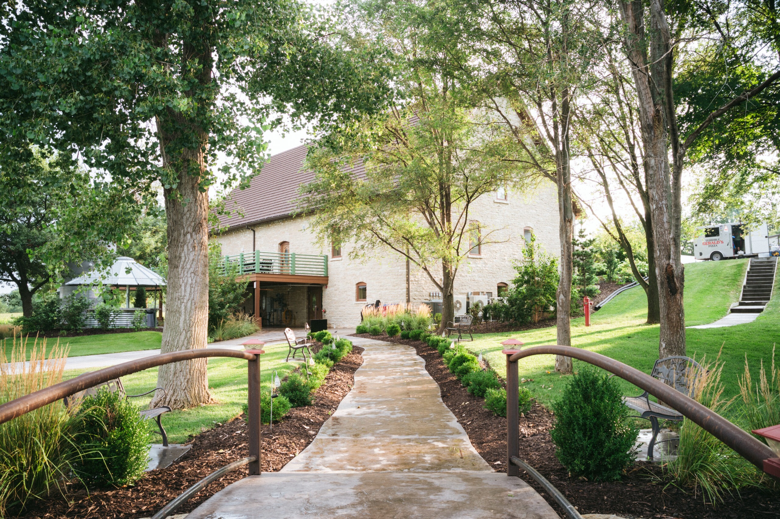 Stone building wedding venue with tree-lined walkway and green railing, surrounded by landscaped gardens