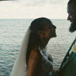 Bride and groom facing each other by ocean window, groom in teal suit with boutonniere