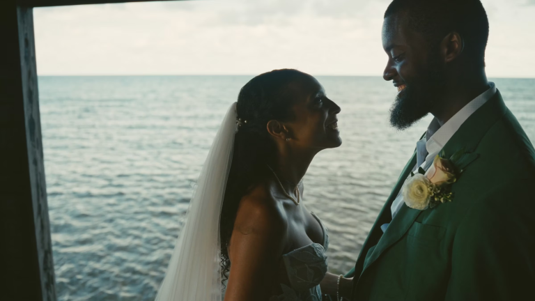 Bride and groom facing each other by ocean window, groom in teal suit with boutonniere
