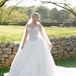 Bride in strapless ball gown with cathedral train standing by stone wall in sunlit field