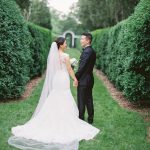 Bride and groom holding hands on manicured garden path lined with tall hedges leading to arched gateway