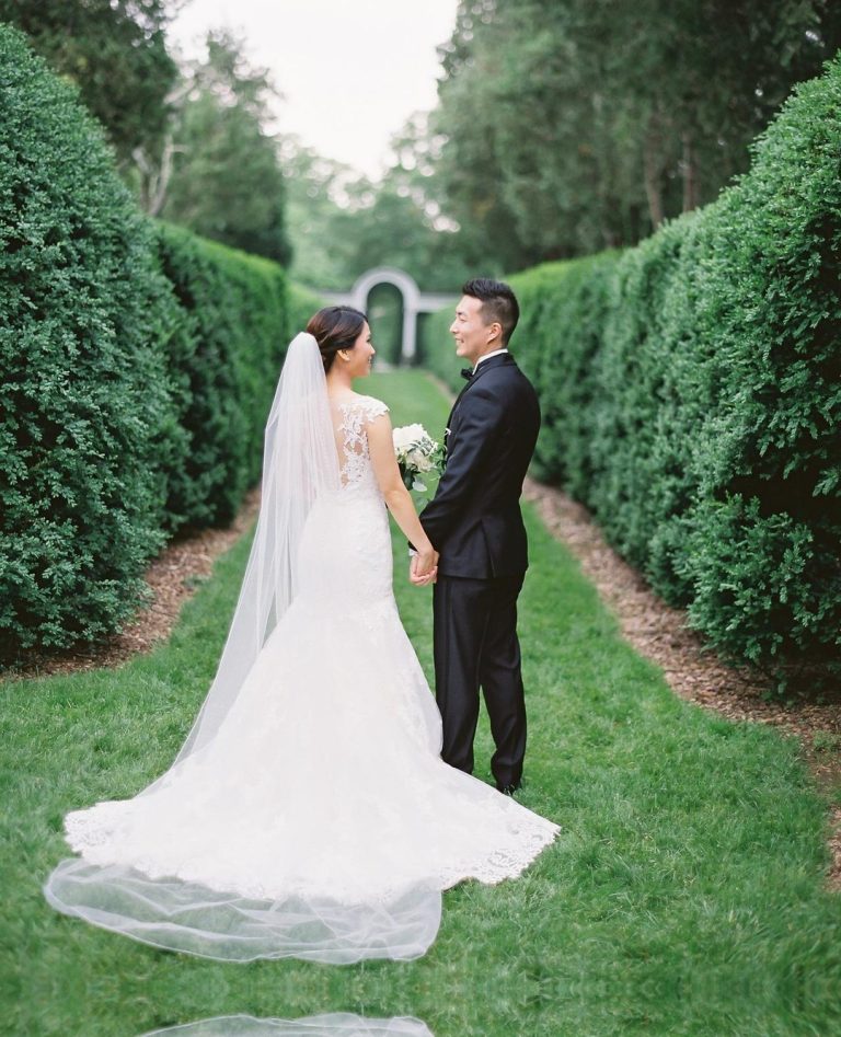 Bride and groom holding hands on manicured garden path lined with tall hedges leading to arched gateway