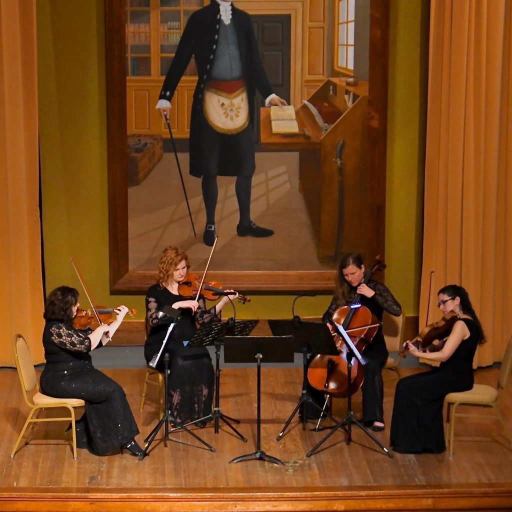 String quartet performing in formal hall beneath large historical portrait of man in Masonic apron