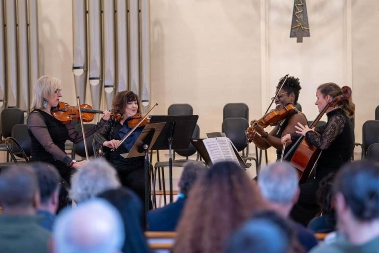 String quartet performing for seated audience in concert hall with organ pipes visible