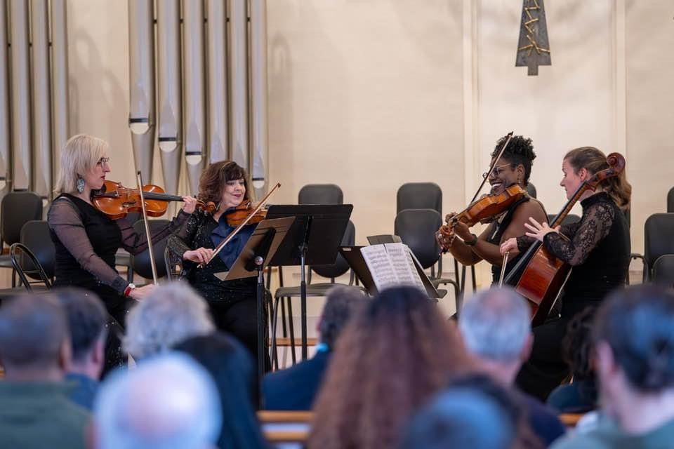 String quartet performing for seated audience in concert hall with organ pipes visible