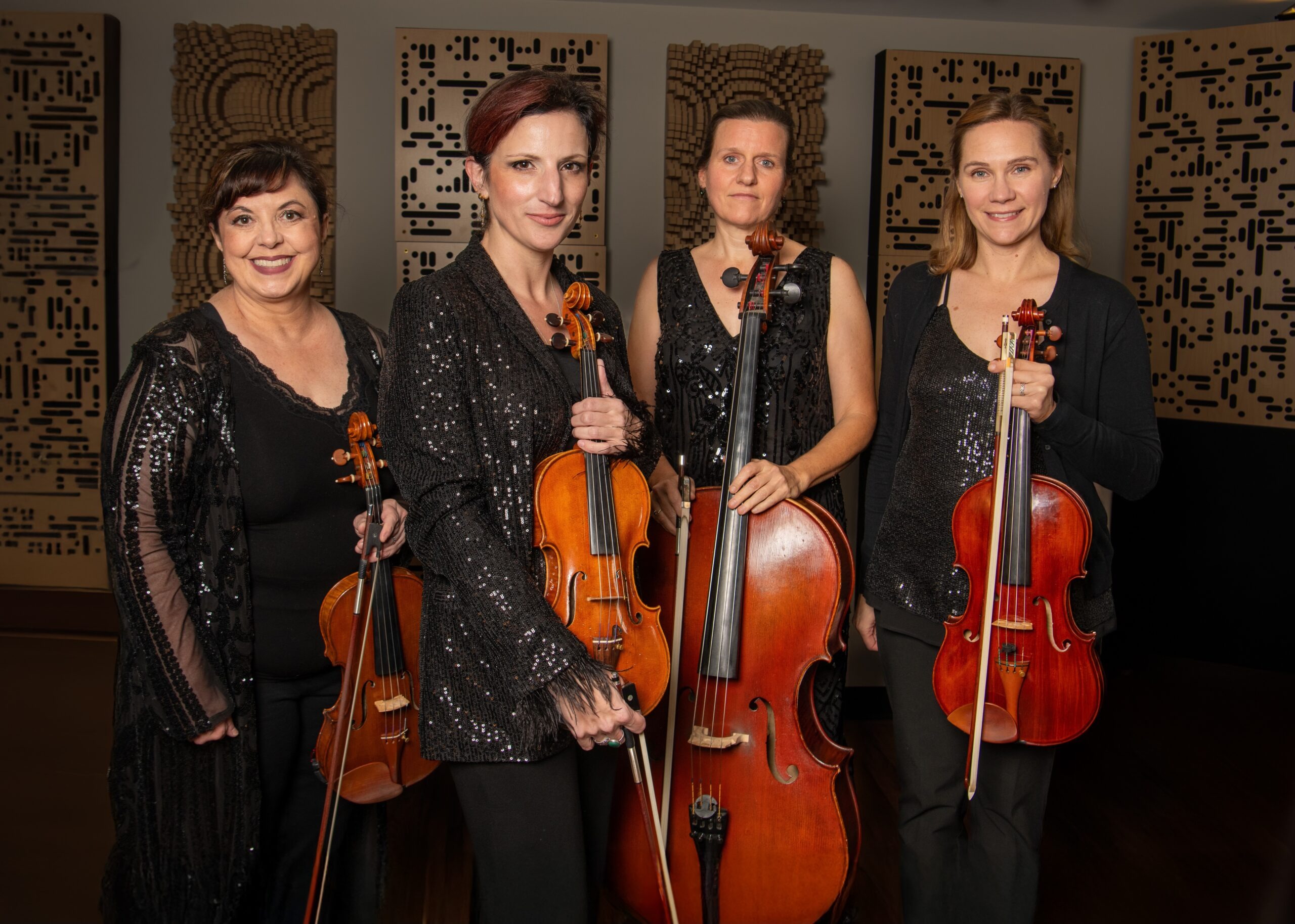 Four female musicians in black evening wear posing with violins and cello against decorative panel backdrop