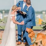 Outdoor wedding ceremony with bride and groom holding hands while two golden retrievers wearing eucalyptus garlands stand between them