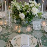 Elegant wedding table setting with white and green floral centerpiece, pillar candles in glass hurricanes, patterned china, and green damask linens
