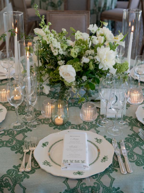 Elegant wedding table setting with white and green floral centerpiece, pillar candles in glass hurricanes, patterned china, and green damask linens
