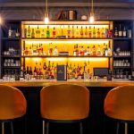 Sophisticated bar with backlit shelves of spirits, pendant lighting, and orange velvet seating
