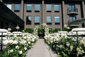 White rose garden ceremony setup with floral aisle borders leading to modern building courtyard