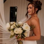Bride with elegant updo hairstyle holding white and cream rose bouquet in rustic venue