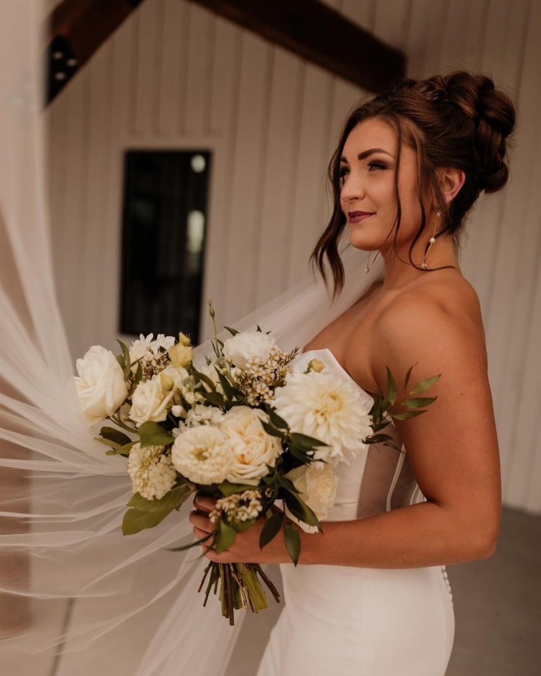 Bride with elegant updo hairstyle holding white and cream rose bouquet in rustic venue