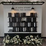 Modern wedding seating chart styled as bookshelf with "A Love Story for the Books" sign, featuring white floral arrangement at base