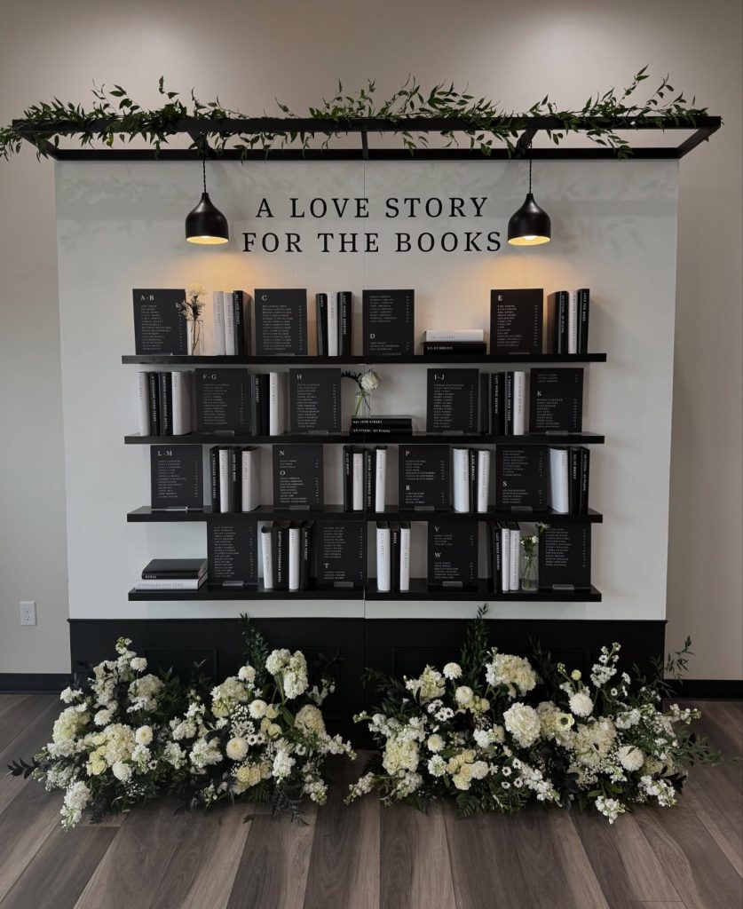 Modern wedding seating chart styled as bookshelf with "A Love Story for the Books" sign, featuring white floral arrangement at base