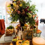 Multi-tiered charcuterie table with dramatic floral centerpiece of yellow, burgundy, and peach blooms above cheese and fruit displays