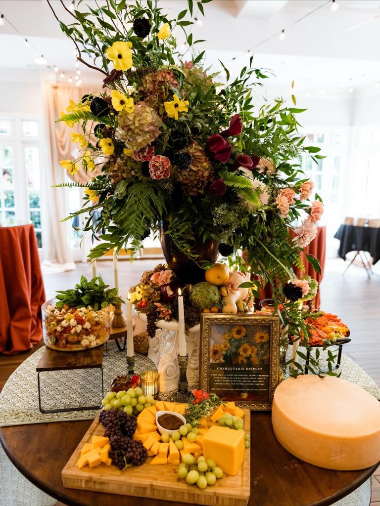 Multi-tiered charcuterie table with dramatic floral centerpiece of yellow, burgundy, and peach blooms above cheese and fruit displays