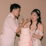 Couple in white attire feeding each other cake from three-tier wedding cake adorned with cascading pink flowers