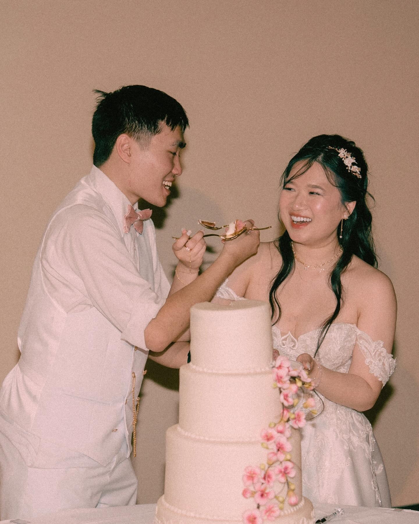 Couple in white attire feeding each other cake from three-tier wedding cake adorned with cascading pink flowers