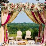 Elaborate mandap ceremony structure with pink, gold, and burgundy drapes, abundant florals, and white chairs on outdoor terrace