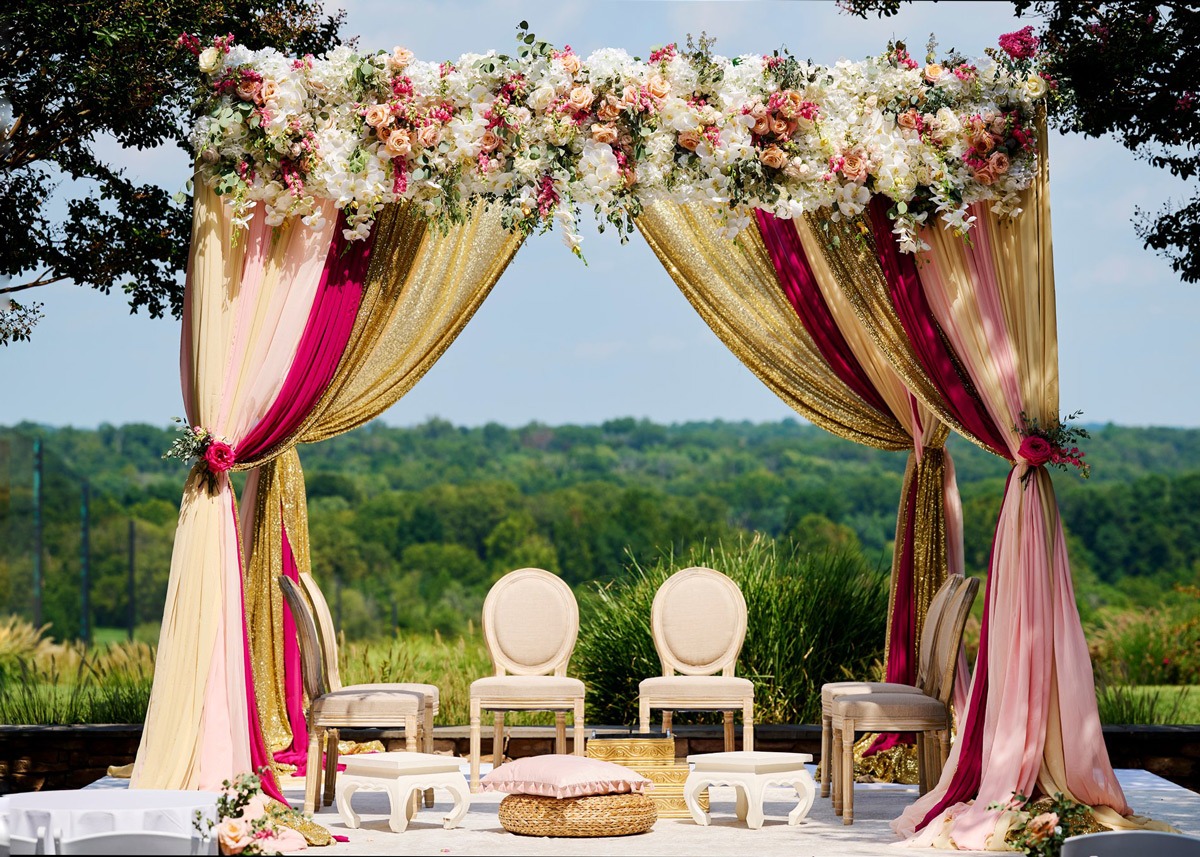 Elaborate mandap ceremony structure with pink, gold, and burgundy drapes, abundant florals, and white chairs on outdoor terrace