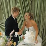 Bride and groom cutting elegant white tiered wedding cake decorated with floral details against sage green drapes