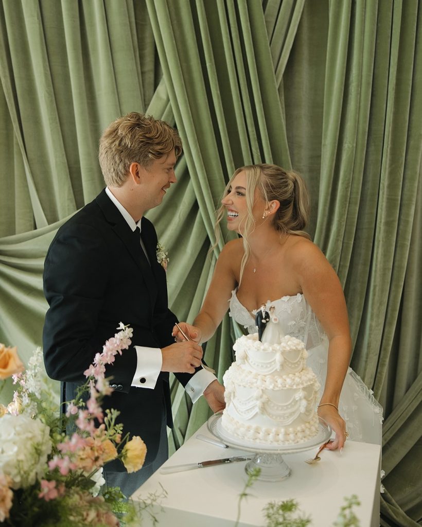 Bride and groom cutting elegant white tiered wedding cake decorated with floral details against sage green drapes