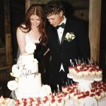 Bride and groom cutting white wedding cake with gold calligraphy topper and fresh flower decorations alongside strawberry dessert bar